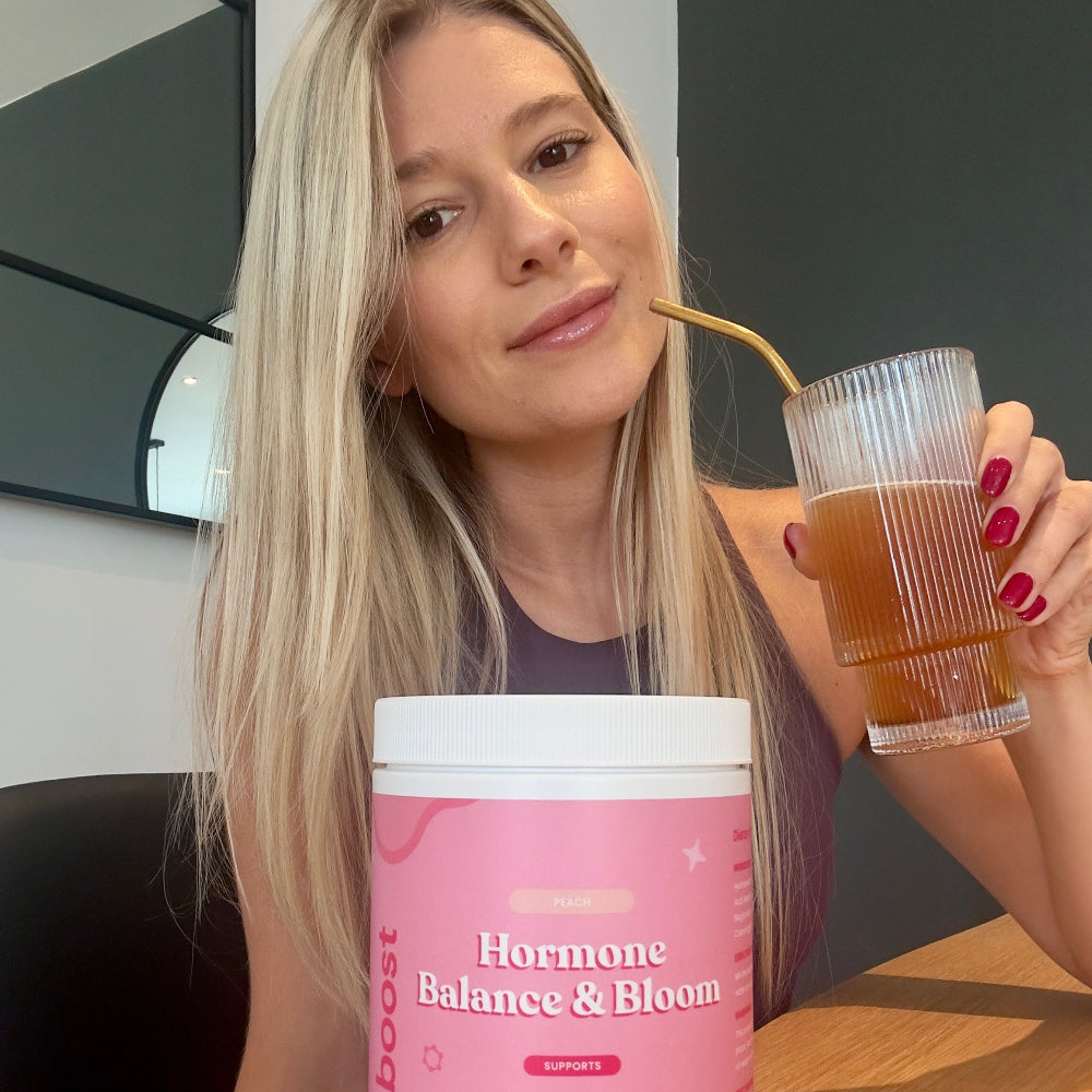 Woman holding a glass of a drink and a container labeled 'Hormone Balance & Bloom' indoors.