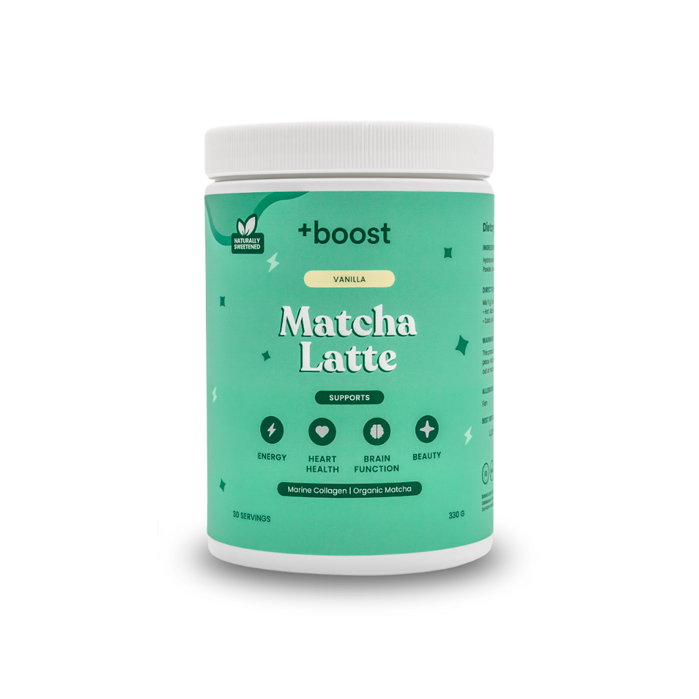 Creamy +boost Matcha Latte in a glass mug with frothy top, featuring vibrant green matcha powder, on a wooden surface.